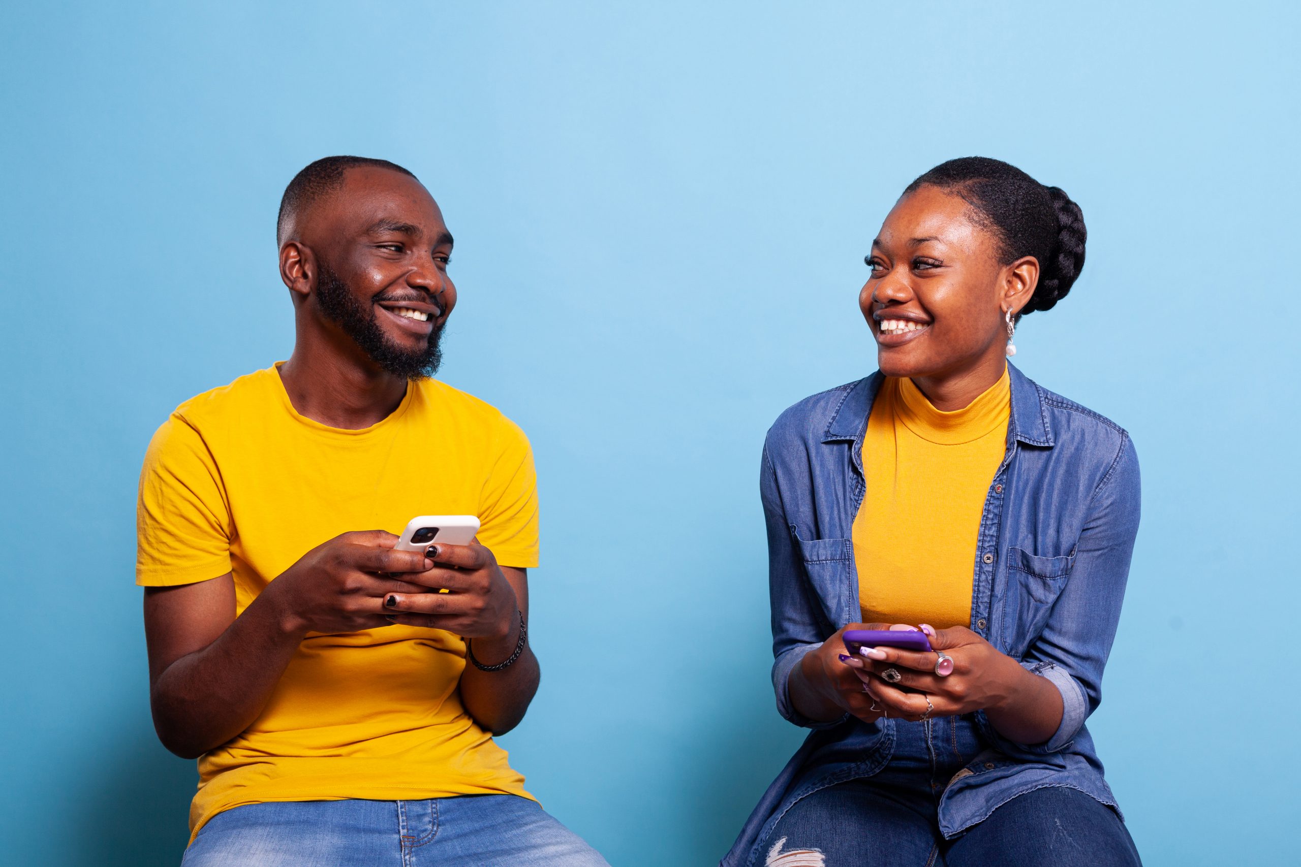 Man and woman holding smartphones and smiling at each other, using mobile phones with touch screen to browse internet network. Modern couple laughing and using social media connection in studio.