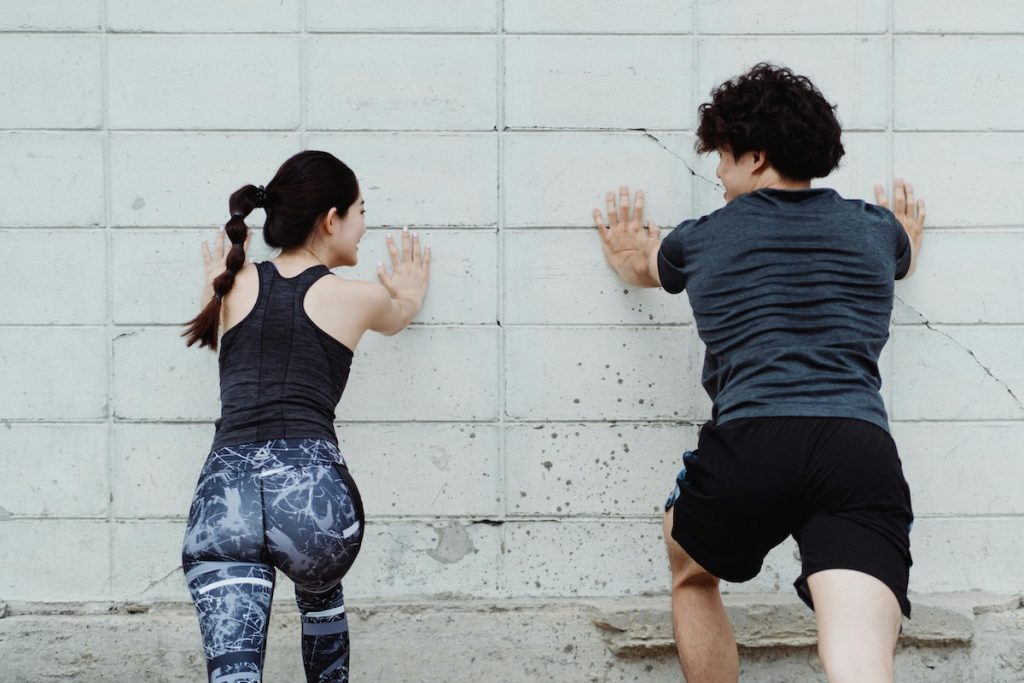 Rear view of a man and woman in sportswear stretching or doing wall push-ups against a white cinder block wall.