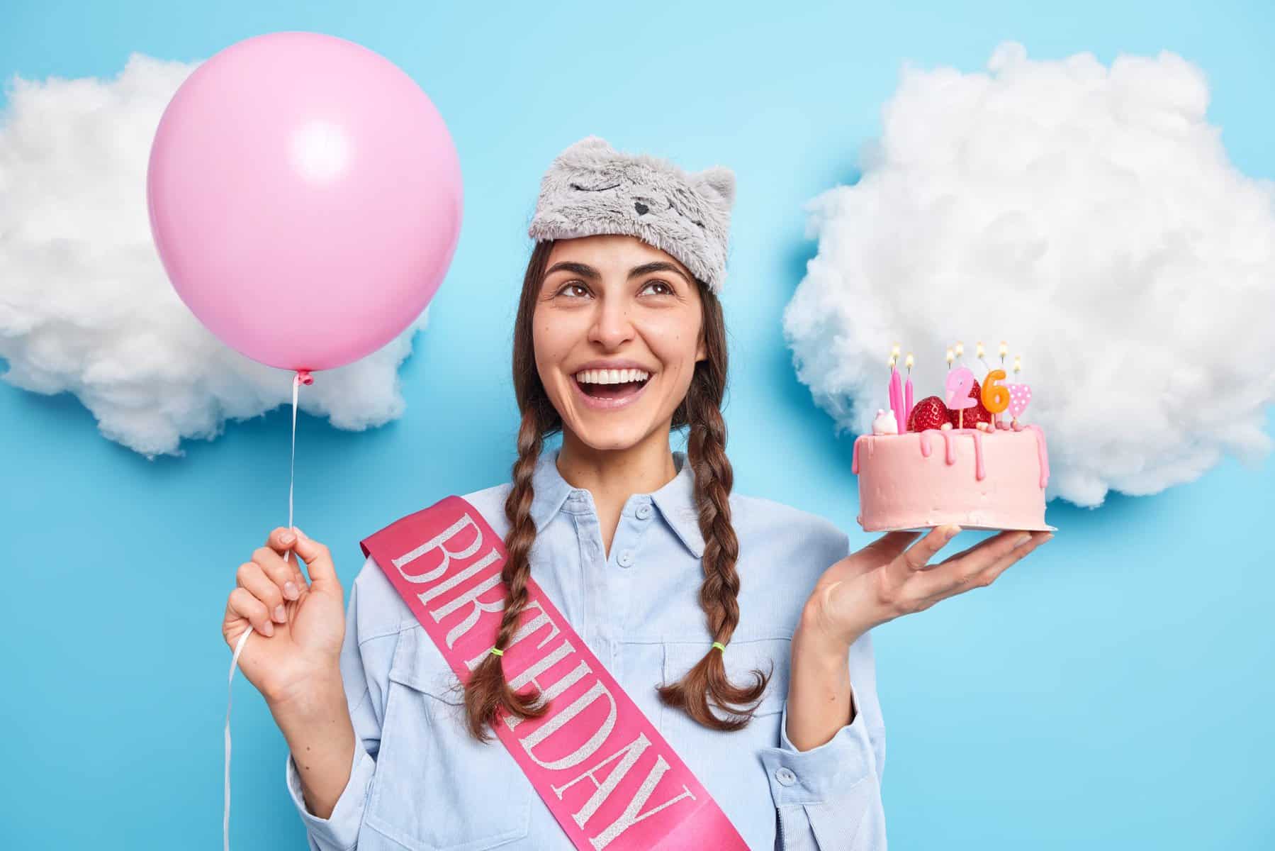 Positive European girl with two pigtails concentrated above has cheerful expression holds festive cake and helium balloon celebrates 26th birthday waits for friends on party accepts birthday congratulations