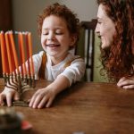 A smiling woman and a young child with curly red hair look happily at a menorah filled with orange candles on a wooden table.