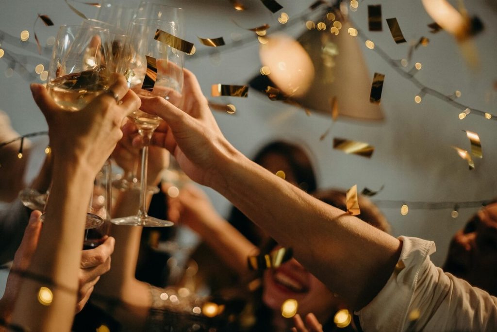 Hands raising glasses in a celebratory toast with golden confetti falling in the air at a festive gathering.