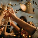 Hands raising glasses in a celebratory toast with golden confetti falling in the air at a festive gathering.