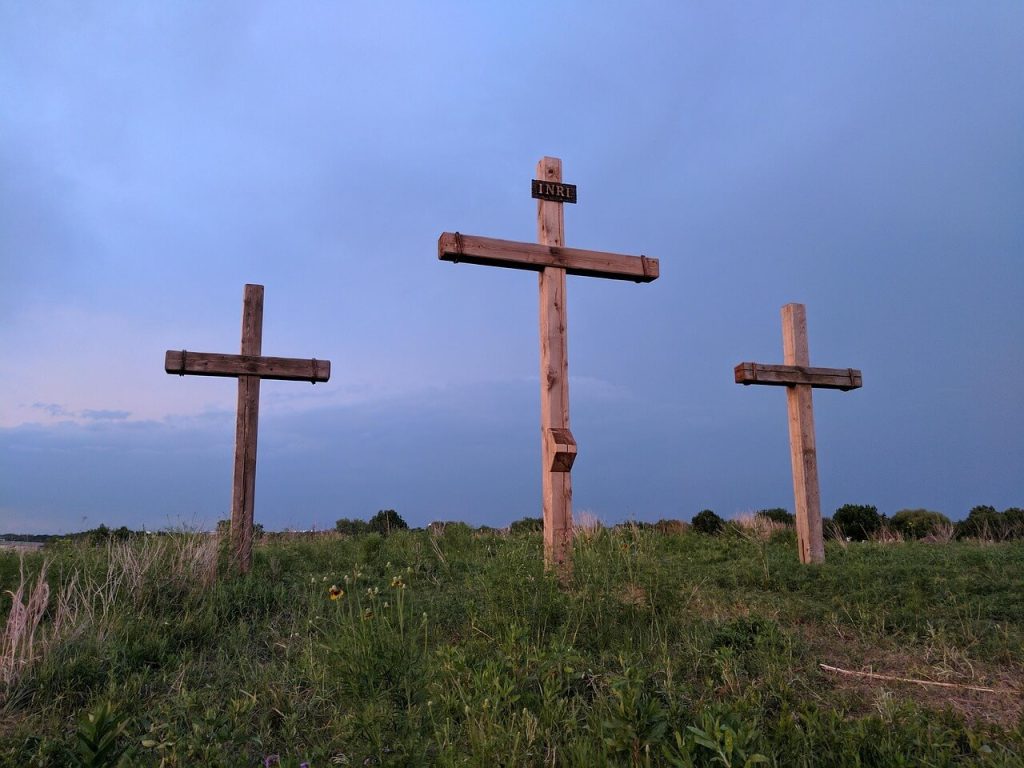 Three wooden crosses standing on a grassy hill under a soft blue and pink evening sky, with the center cross labeled ‘INRI.’