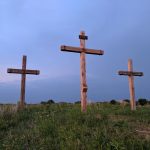 Three wooden crosses standing on a grassy hill under a soft blue and pink evening sky, with the center cross labeled ‘INRI.’