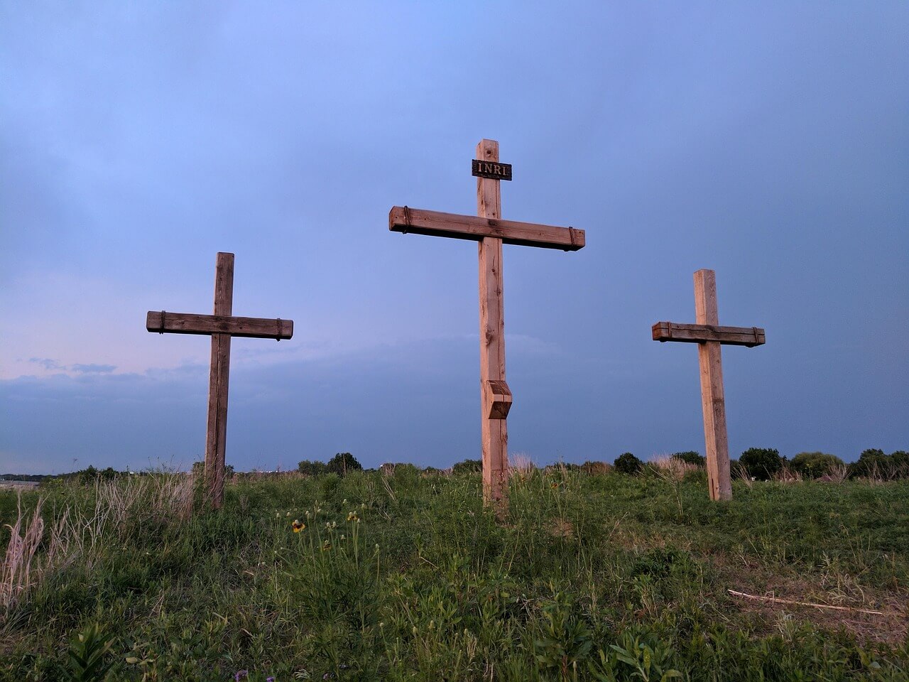 Three wooden crosses standing on a grassy hill under a soft blue and pink evening sky, with the center cross labeled ‘INRI.’